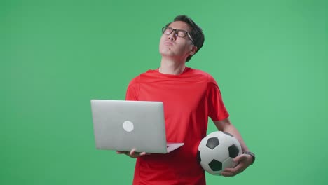 un hombre asiático con una pelota se arrepiente de animar al fútbol en una computadora portátil en el estudio de pantalla verde