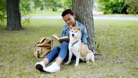 cheerful mixed race girl dog owner is reading book and stroking her cute puppy sitting on grass in park on summer day. backpack, green lawn and trees are visible.