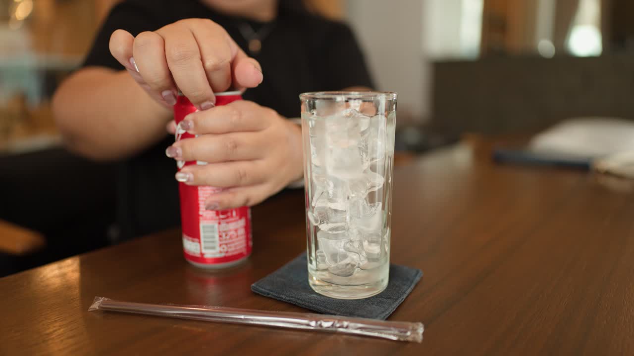 Person pours cola from can into tall glass of ice in casual dining setting
