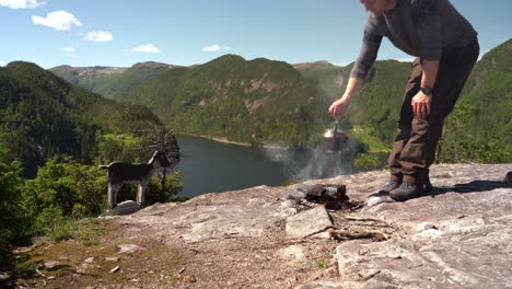 Man-removing-stainless-steel-kettle-with-freshly-brewed-coffee-from-bonfire---Caucasian-male-with-beautiful-Norway-mountain-and-fjord-scenery-background