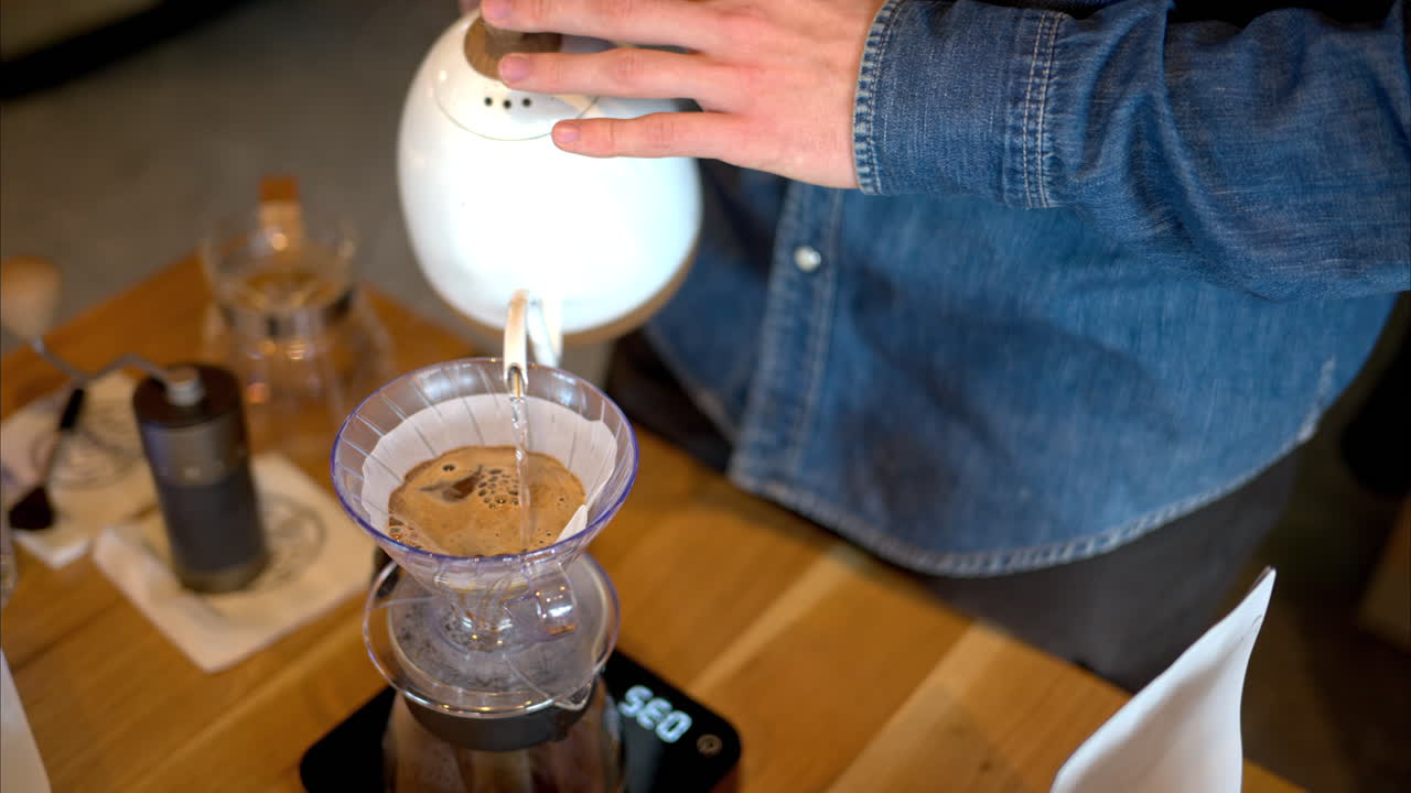 Close up of a man brewing pour-over drip coffee in a slow, circular motion on a wooden table