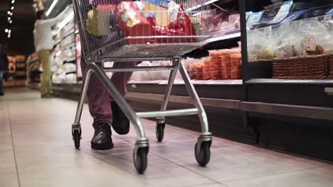 Shopping-in-the-supermarket,-view-of-the-male-legs-from-inside-the-trolley.-A-guy-pushing-full-of-goods-shopping-trolley-by-the-supermarket-aisle.-Cropped,-close-up-footage