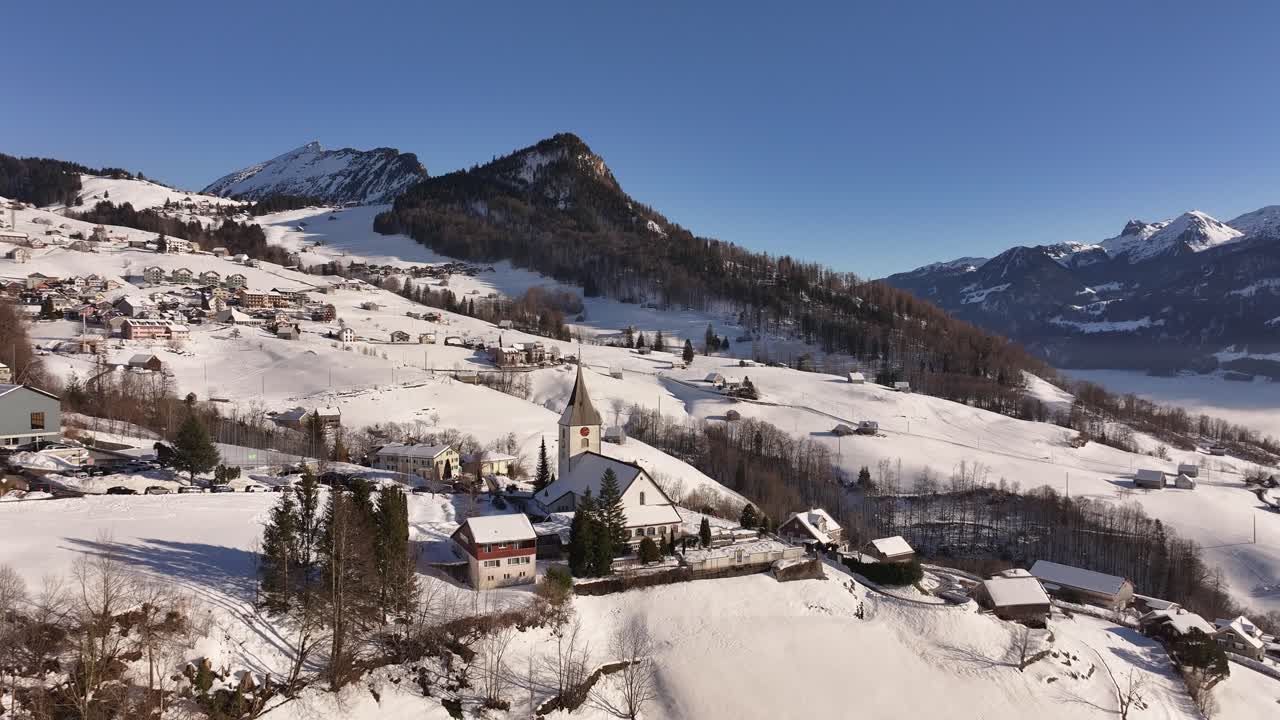 Scenic aerial view of Amden, Switzerland covered in fresh snow, stunning mountain backdrop, charming village houses and picturesque church.