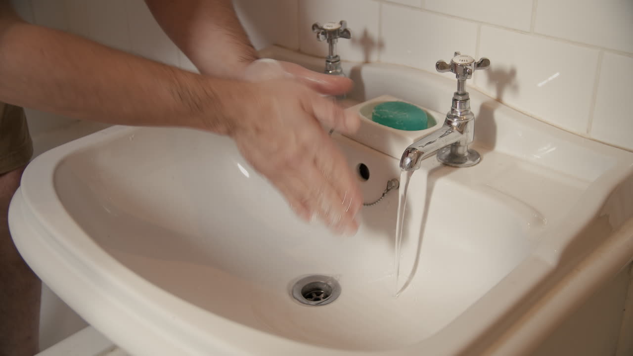 Male hands wash and scrub with green soap for twenty seconds duration in white bathroom sink. Three quarters angle, interior, day light.