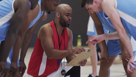 equipo de baloncesto masculino diverso y entrenador con clipboard en discusión en la cancha cubierta, en cámara lenta