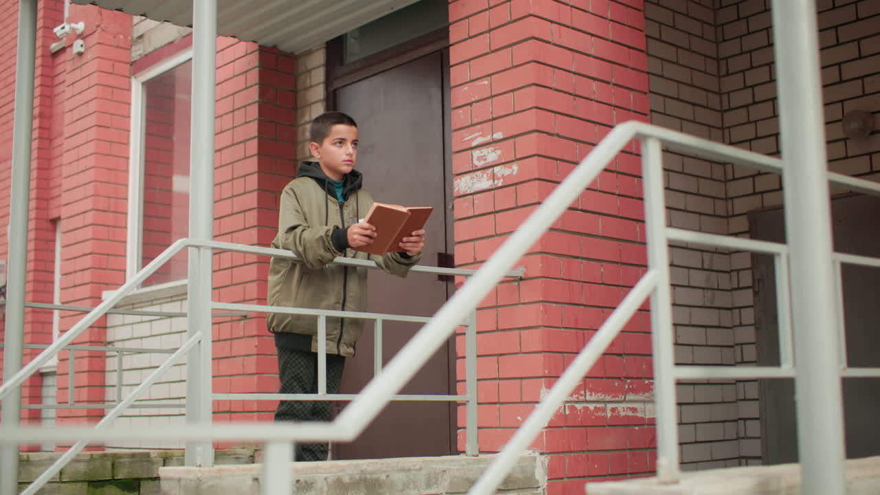 Adolescent in hooded jacket stands near red brick building entrance reading brown book with focus, leaning on railing outdoors while showing serious concentrated expression in quiet environment