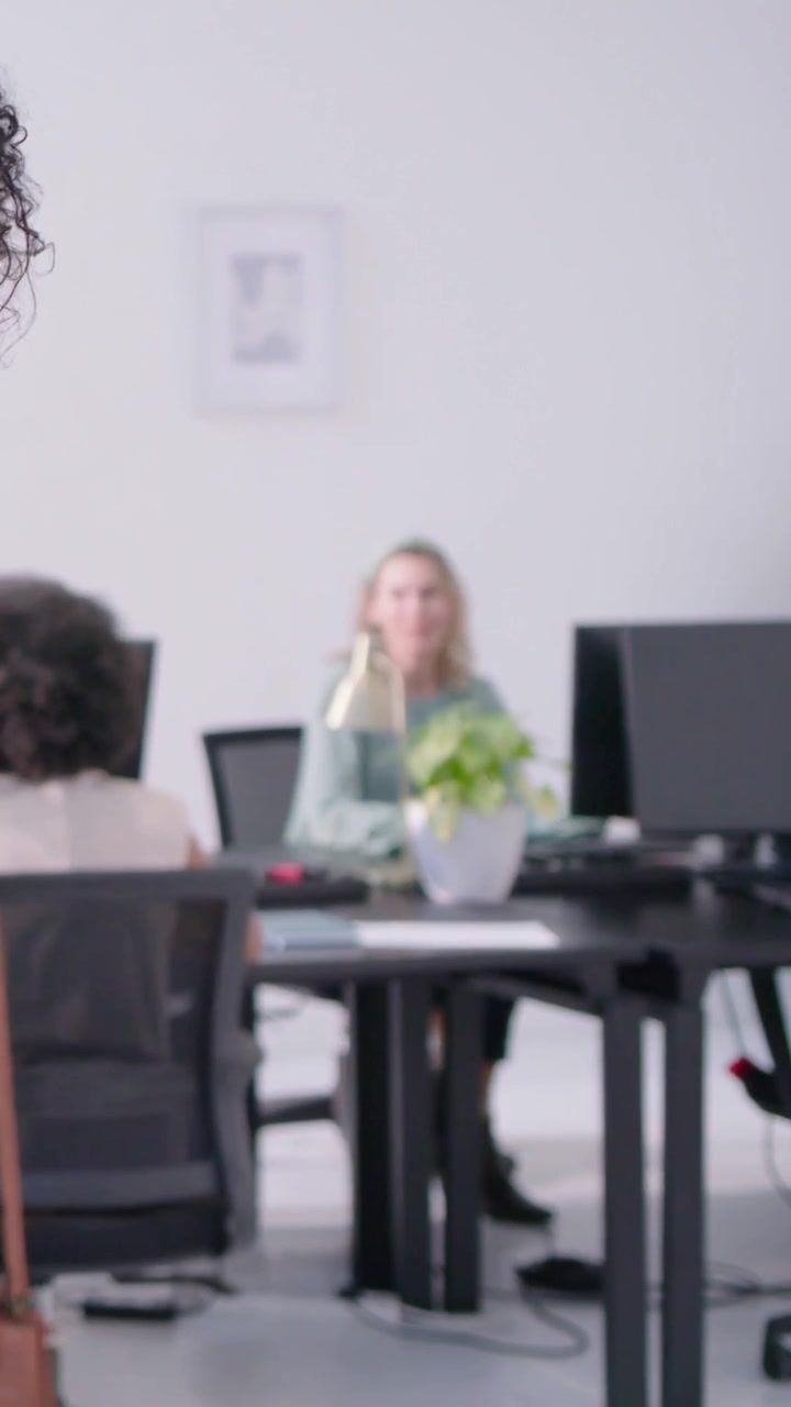 Video verticale: Bionda sorridente con un top verde, appoggiata in ufficio, che reagisce all'ingresso di un collega, monitor.