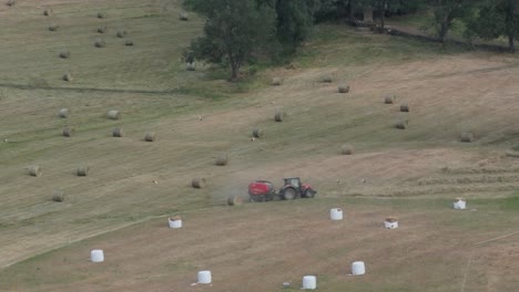 167mm-drone-footage-of-a-meadow-being-harvested-by-two-agricultural-machines,-one-puts-the-hay-into-rows-and-the-other-collects-it-and-makes-round-alpacas