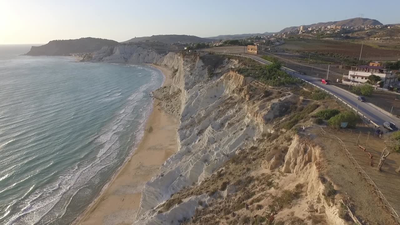 Breed luchtpanorama van de witte kliffen van Scala dei Turchi en het zandstrand langs de turquoise Middellandse Zee, Sicilië, Italië, met kustweg, heuveldorpjes en verre heuvels bij zonsondergang.