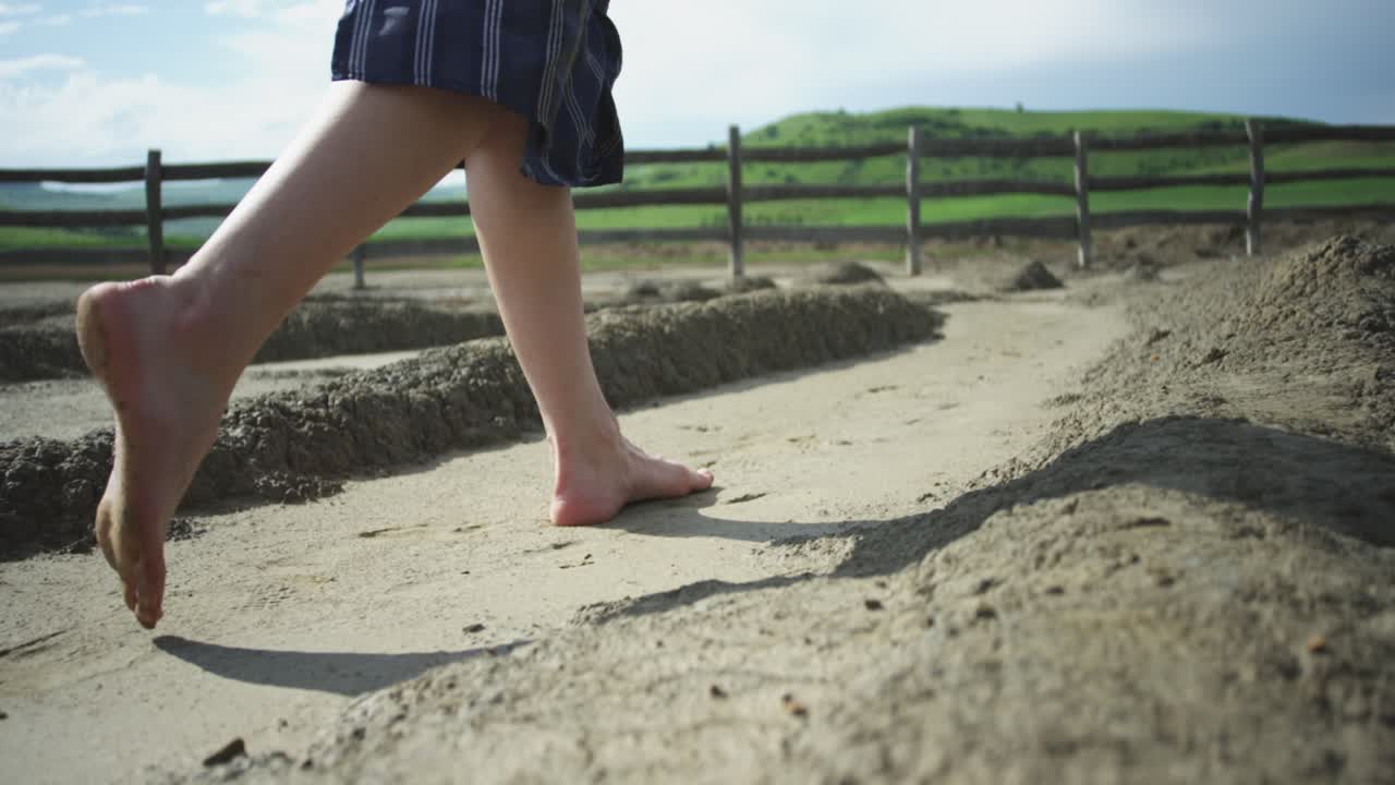 A low-angle tracking shot follows a woman's bare feet as she walks along a path of dry, cracked earth at the Mud Volcanoes. A feeling of freedom and discovery.