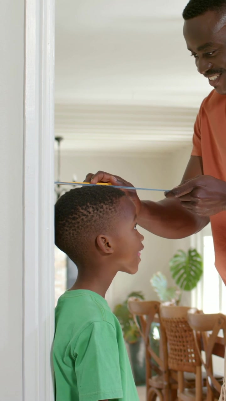 Vertical video: Seeing ruler, dad orange tee measuring son in green at home, using yellow pencil