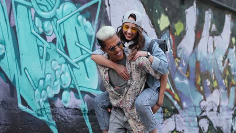 un joven que le da a su novia un piggyback cerca de una pared de graffity en la calle