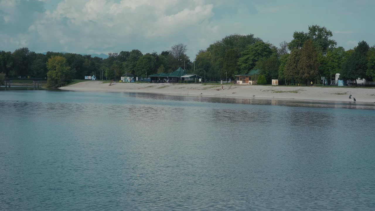 Ampia inquadratura della spiaggia e delle strutture sul lungomare del lago Jarun a Zagabria, Croazia. L'acqua calma riflette il cielo, con persone che si rilassano sulla riva ed edifici visibili sullo sfondo.