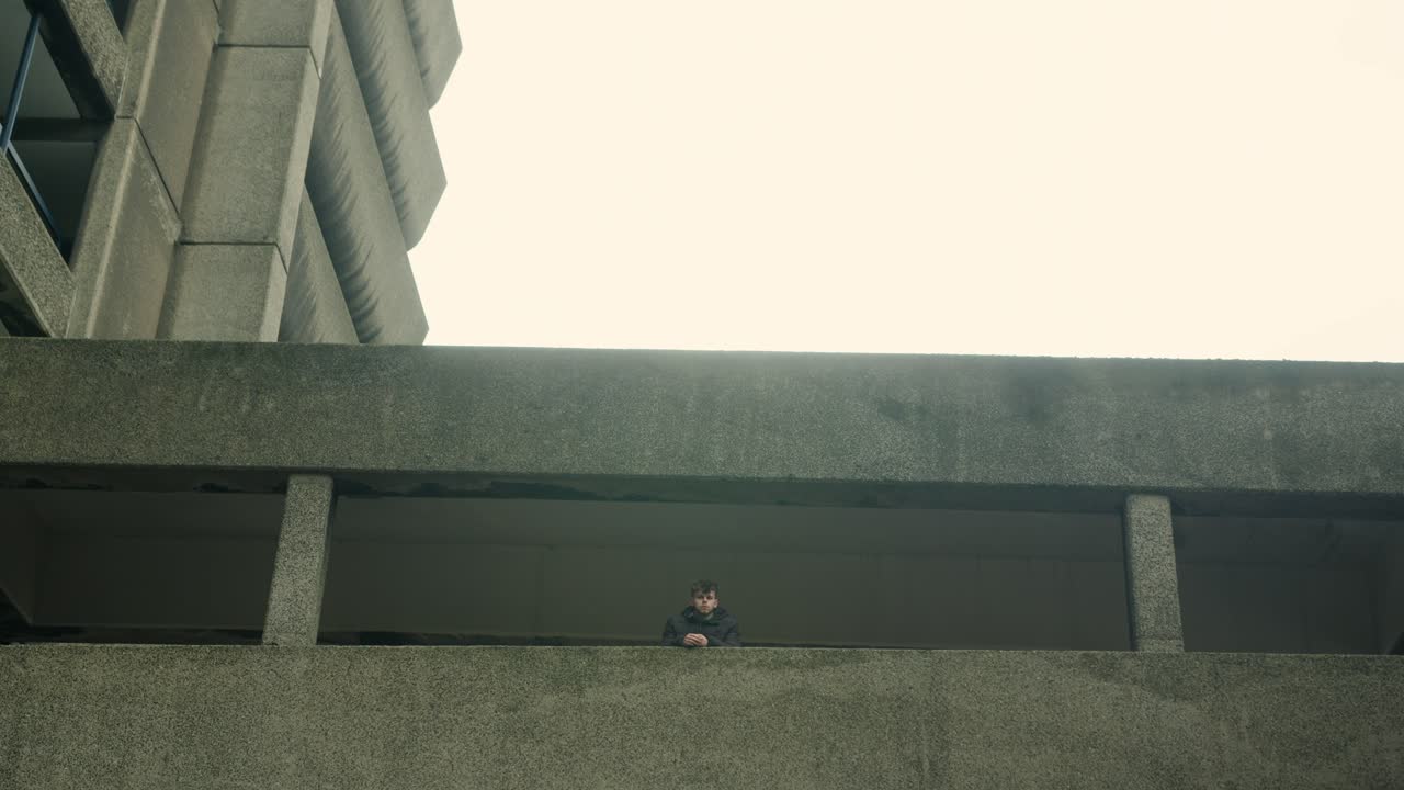 Man stares off into distance leaning against concrete walkway under heavy Barbican housing structure, angled framing with moody light