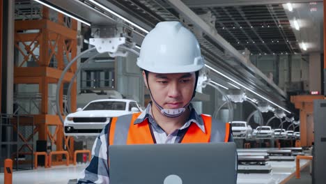primer plano de un ingeniero masculino asiático con casco de seguridad trabajando en una computadora portátil mientras estaba de pie en una fábrica de automóviles ev