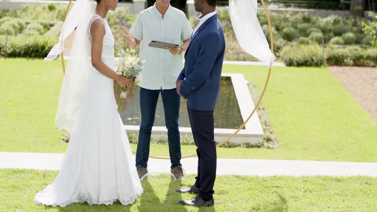 hombre biracial oficiando el matrimonio de una feliz pareja afroamericana en un jardín soleado, en cámara lenta