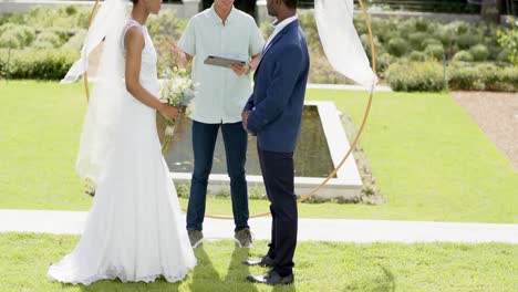 biracial man officiating marriage of happy african american couple in sunny garden, in slow motion