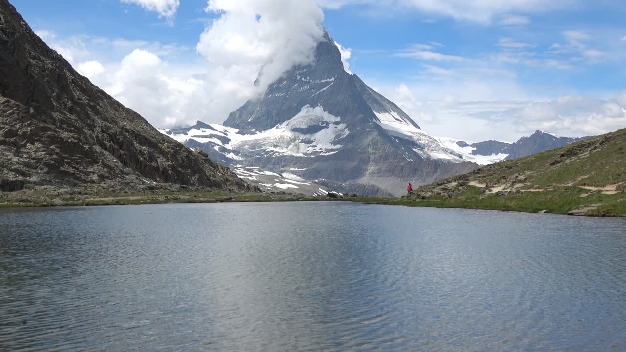 Ampia inquadratura di un lago di montagna calmo con il Cervino che si erge sopra creste innevate e nuvole, filmato vicino a Zermatt, Vallese, Svizzera, in una luminosa giornata estiva