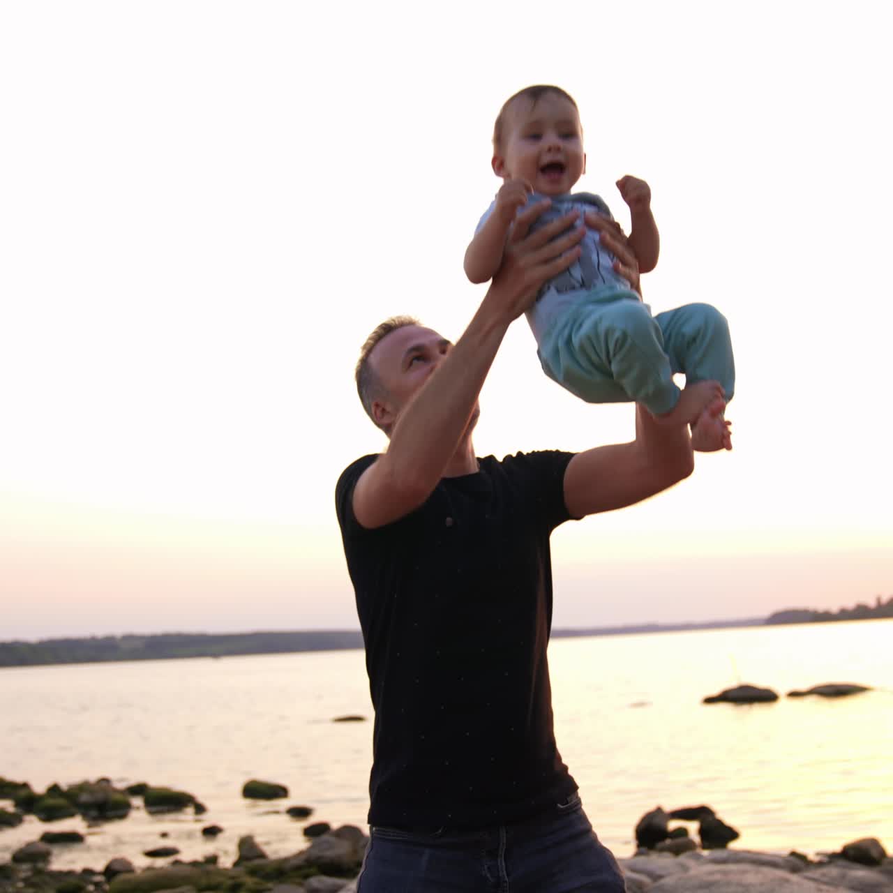 Un adorabile bambino felice viene lanciato in aria dal suo papà. Padre e figlio si divertono vicino al fiume al tramonto.