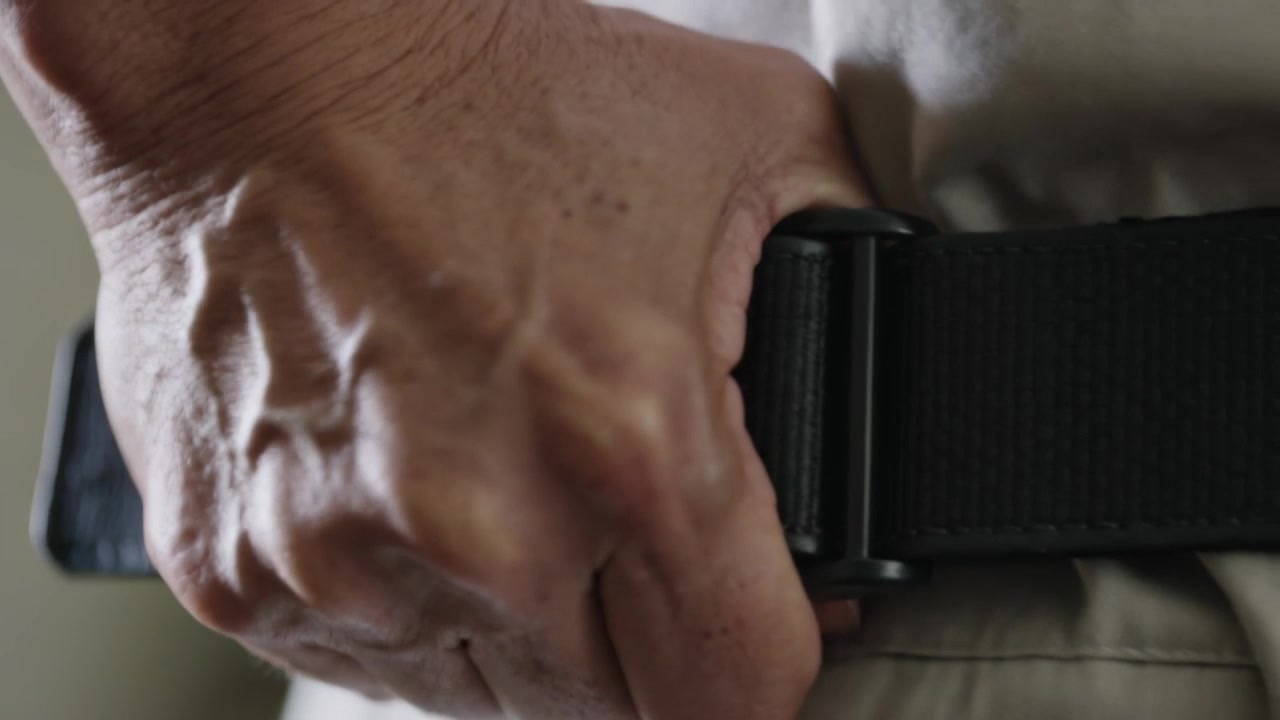 Entering right-hand pulling webbing-belt into buckle on khaki pants waist in room, securing holster