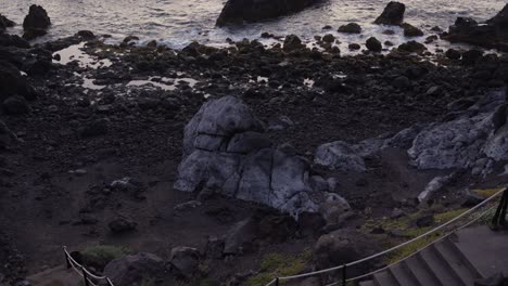 Panning-down-from-ocean-to-dark-volcanic-coast,-revealing-concrete-stairs