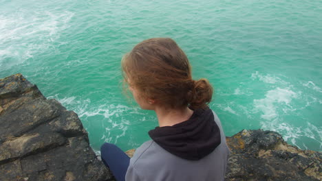 Caucasian-Teenage-Boy-Looking-Away-With-A-Sad-Face-While-Sitting-on-Rocky-Cliff-Over-The-Ocean---Overhead-Zoom-out