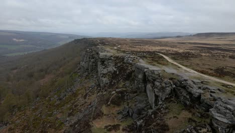 Aerial-drone-footage-descending-over-a-dramatic-rock-formation-in-a-large,-rural-environment-with-open-moorland-and-a-dramatic-cloudy-sky