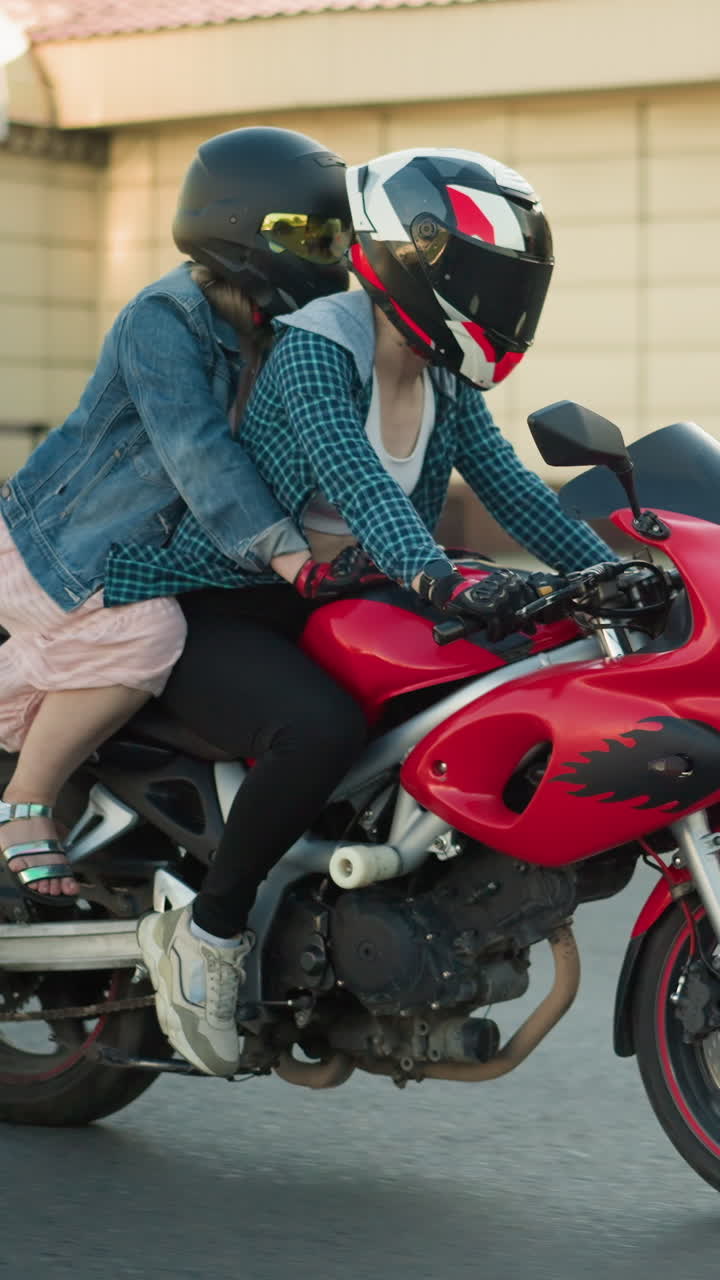 dos mujeres montan una bicicleta eléctrica roja a través de una calle urbana, con la camisa de botón abajo del jinete abierta, pasan por un edificio, con la luz del sol reflejándose en ellos