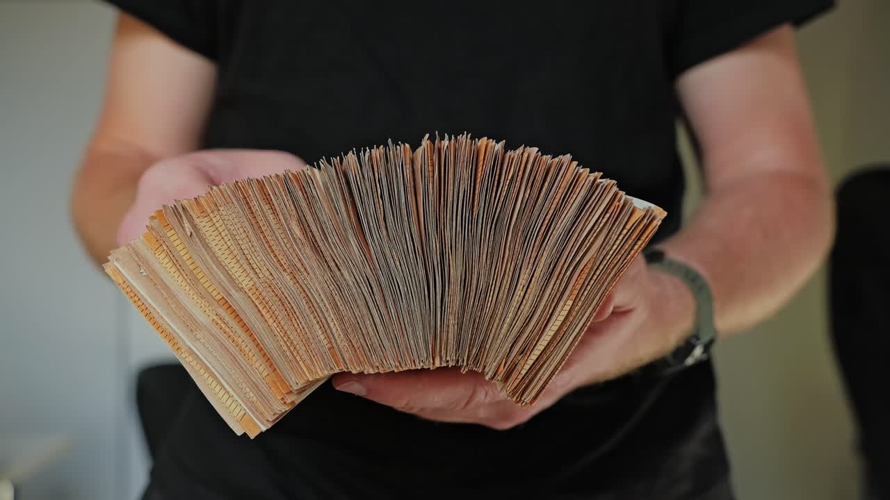 A man in a black shirt slowly opens a large stack of banknotes like an accordion