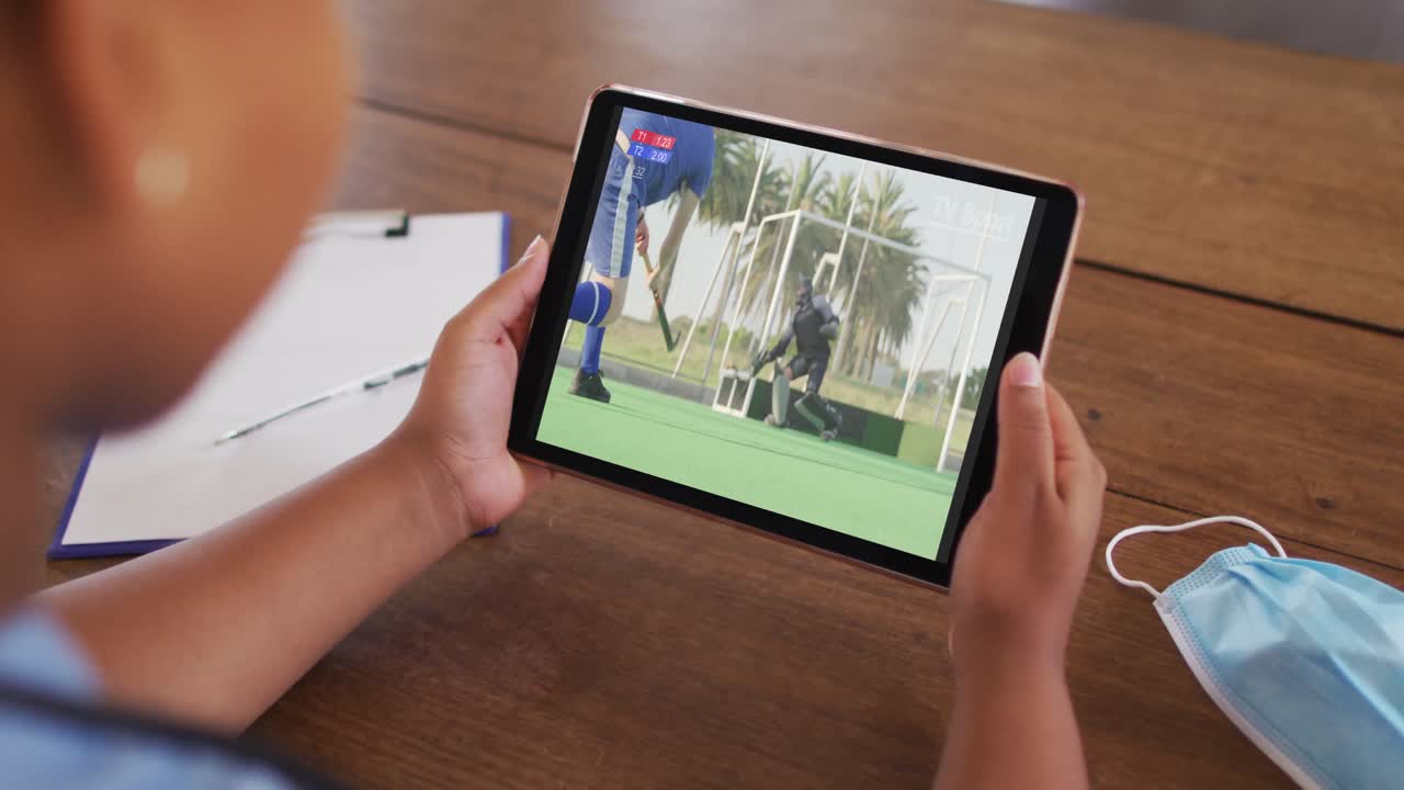 태블릿에서 하키 경기를 시청하는 얼굴 마스크와 함께 테이블에 앉아있는 여성의 복합