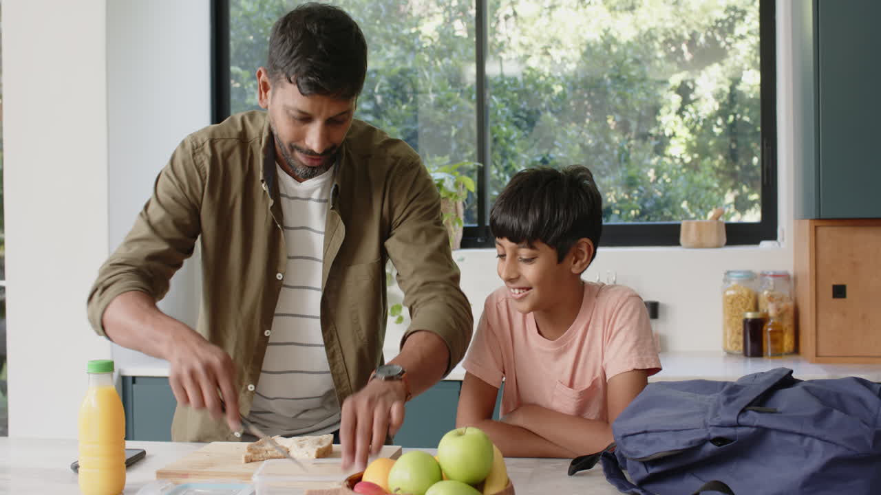 Preparing sandwiches, Indian father and son bonding in kitchen with backpack and fruit