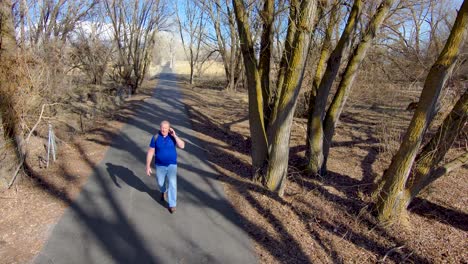 Un-Hombre-Mayor-En-Un-Paseo-Por-La-Naturaleza-No-Puede-Alejarse-De-Su-Smartphone-Y-Habla-Mientras-Camina---Vista-Aérea-En-Cámara-Lenta