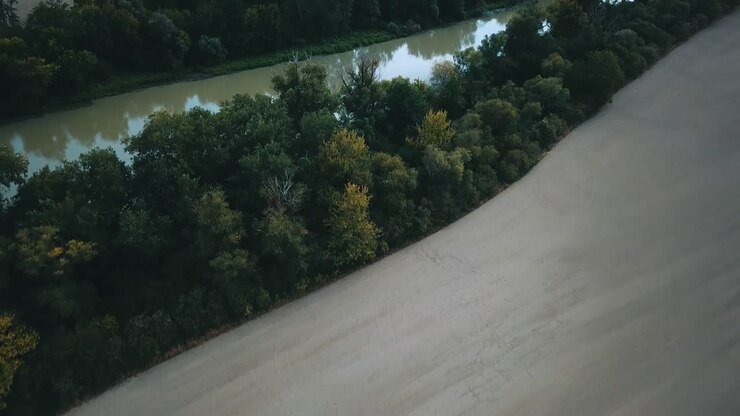 Drone shot over slow muddy river between fields and trees