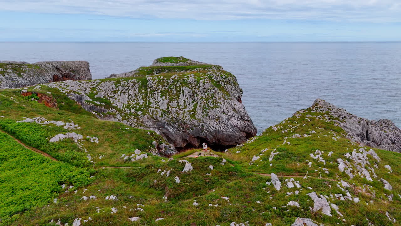 Couple stands at the precipice over the seascape. Drone footage above the rocky shore of Spain, Europe.