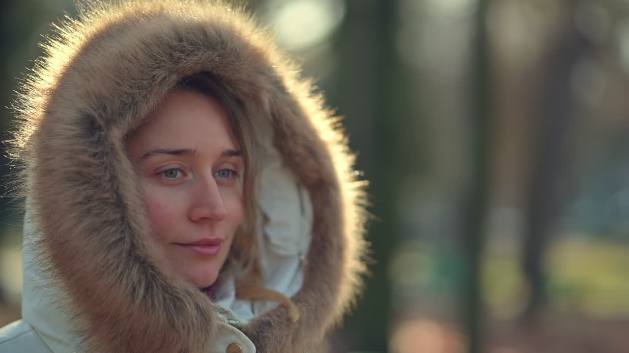 A woman enjoys a peaceful walk in the park on a sunny winter day. She is dressed warmly in a hooded coat, surrounded by trees and soft sunlight