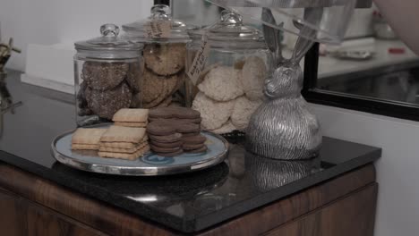 Una-Mujer-Levanta-Una-Pantalla-De-Vidrio-Que-Revela-Varias-Galletas-De-Mantequilla-Y-Coco-En-Una-Pastelería-Artesanal,-4k