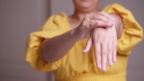 a close-up of a woman wearing a yellow dress is stretching her left arm while pinching and massaging herself from her wrist down to her fingers