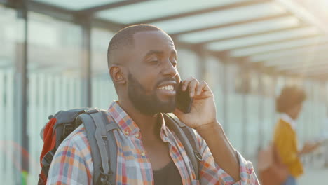 Junger-Gutaussehender-Afrikanisch-amerikanischer-Mann-Mit-Rucksack,-Der-Auf-Dem-Smartphone-Spricht-Und-Am-Bahnhof-Fröhlich-Lächelt