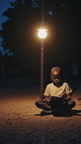 african child sits cross legged on the ground, illuminated by a street lamp, engrossed in his book, providing a heartwarming scene of learning and perseverance