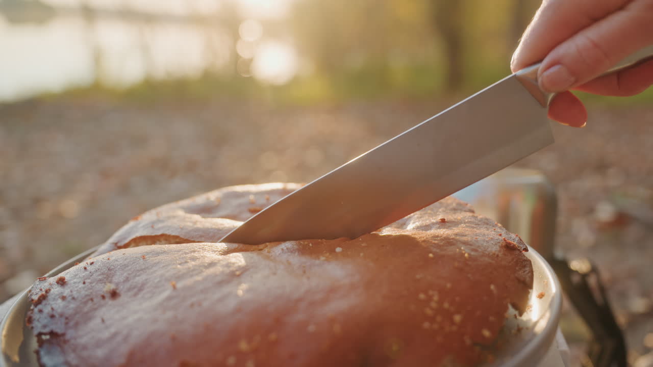 Sunlit camping cake being sliced outdoors, closeup of golden crust glistening with butter, knife pressing into soft crumb on skillet near lake, morning haze and warm bokeh creating cozy rustic
