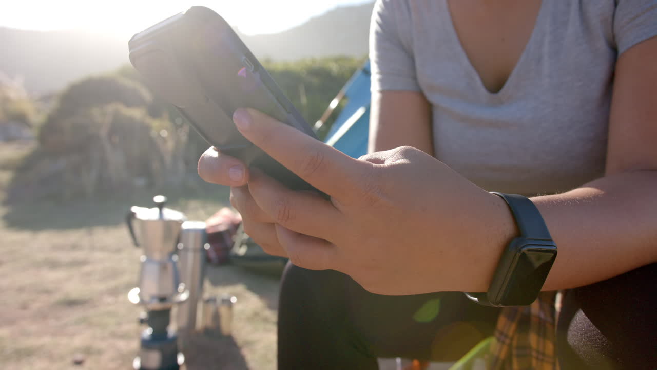 Donna seduta all'aperto con lo smartphone vicino all'attrezzatura da campeggio durante un'escursione in montagna.