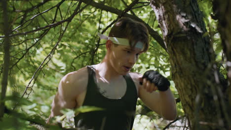 warrior wearing gloves and headband delivers powerful punch to rugged tree branch under leafy canopy, elbow brushing bare twigs as he trains combat technique amid dappled sunlight and dense forest