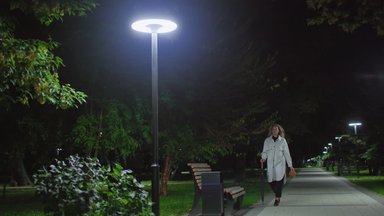 Lady walks calmly toward wooden bench holding umbrella and book under glowing night streetlight surrounded by swaying green leaves, sits down, cross legs, adjust hair, and read