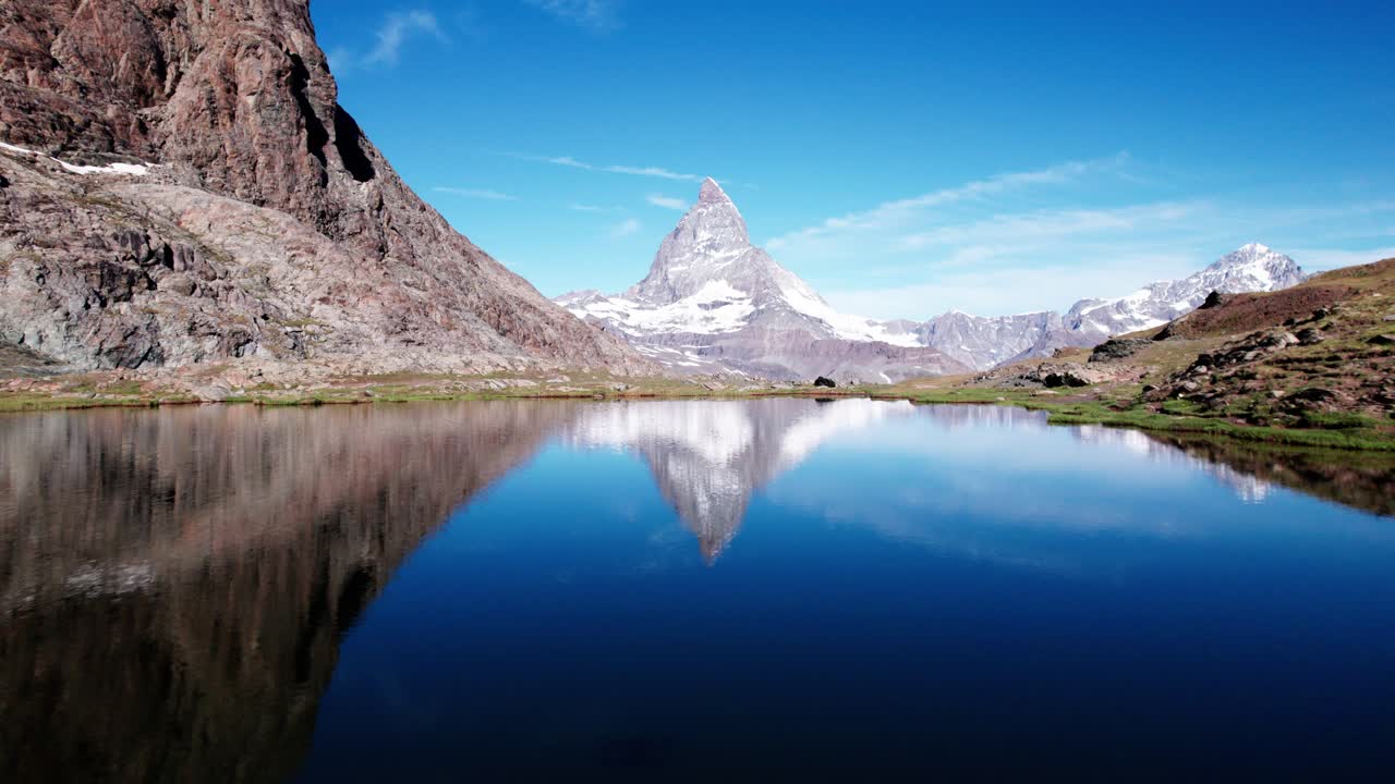 Ripresa 4K da drone del Cervino perfettamente riflesso in un calmo lago alpino, circondato da terreno roccioso e cieli blu tersi, che crea un paesaggio montano svizzero sereno e iconico.