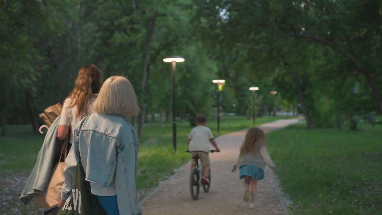 back view of boy riding bike along winding park trail while sister runs after him and mother follows behind carrying art supplies under dappled lamplight creating cozy family activity scene