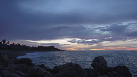 Nightfall-beach-bay-man-enjoys-his-time-looking-at-sunset,-sits-on-rocks
