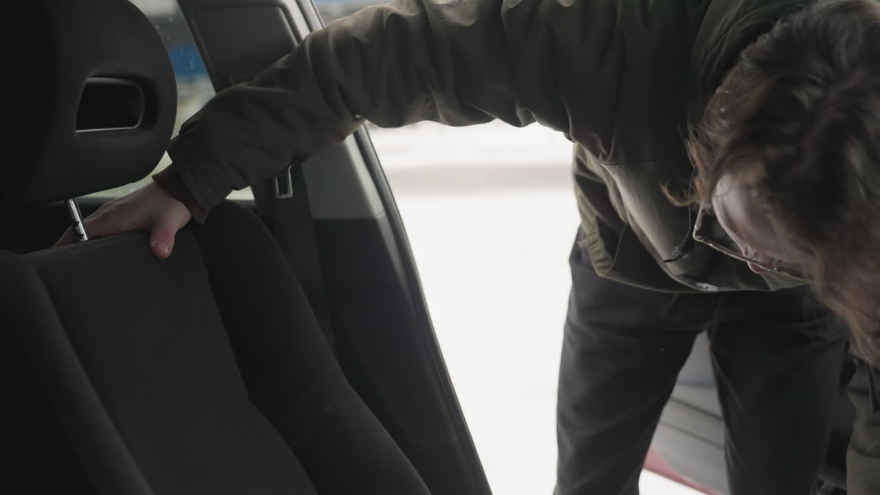close up man in green winter jacket bends forward to adjust driver seat inside car during snowy day with snowy background visible through open door and blurred vehicles in distance