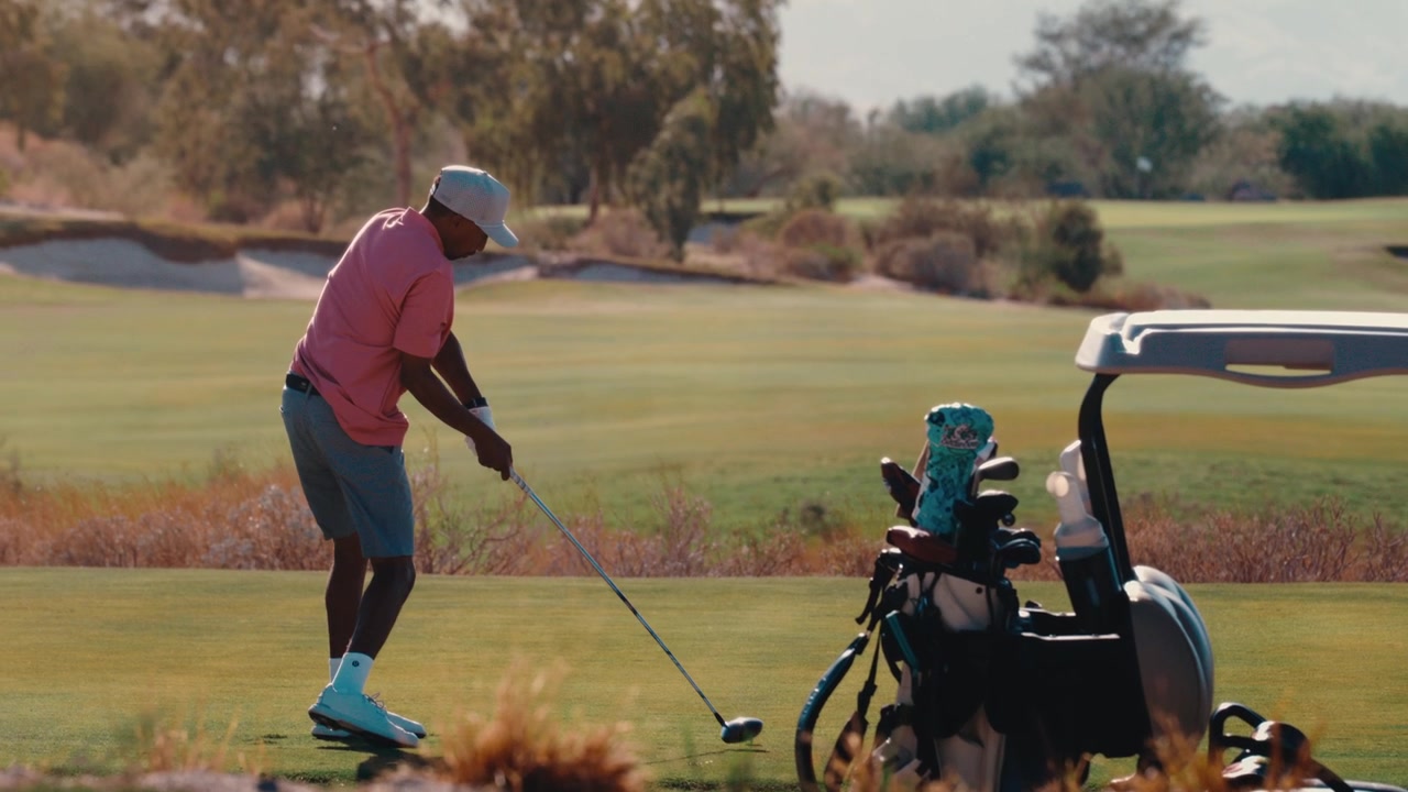 An African American golfer rips a driver off the tee and watches the ball fly toward hazy desert mountains at golden hour, in slow motion. Works for luxury golf and travel ads.