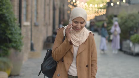 muslim woman wearing hijab walking along city street talking on mobile phone