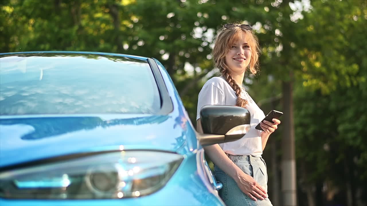 Una giovane donna bionda usa lo smartphone a una stazione di ricarica per auto con un'auto elettrica in carica nelle vicinanze. Chisinau, Moldova. Rallentatore